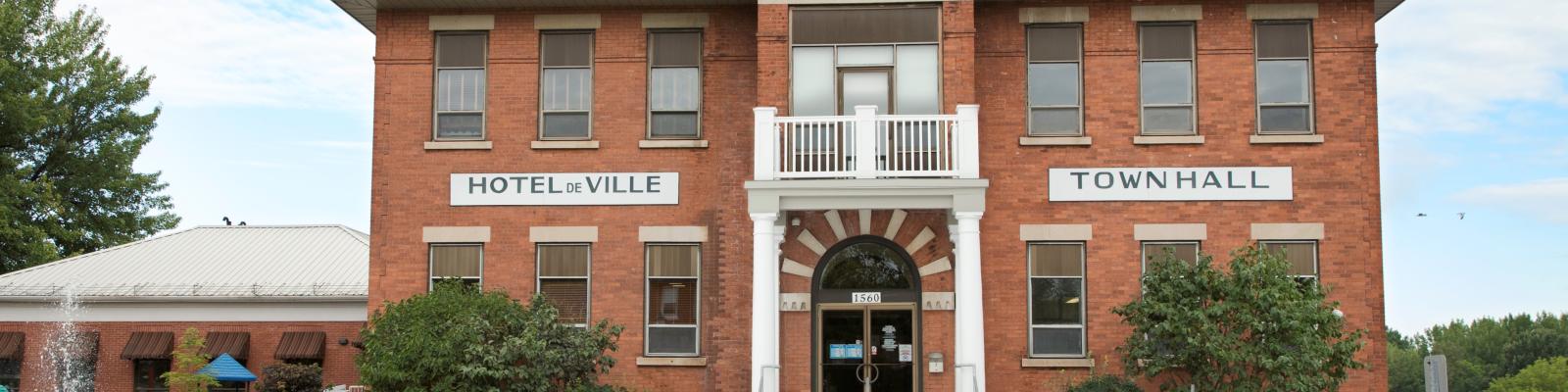 Red brick town hall with white pillars, surrounded by greenery and flowers.