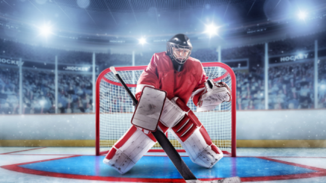 Hockey goalie in red gear guards the net in a packed stadium.