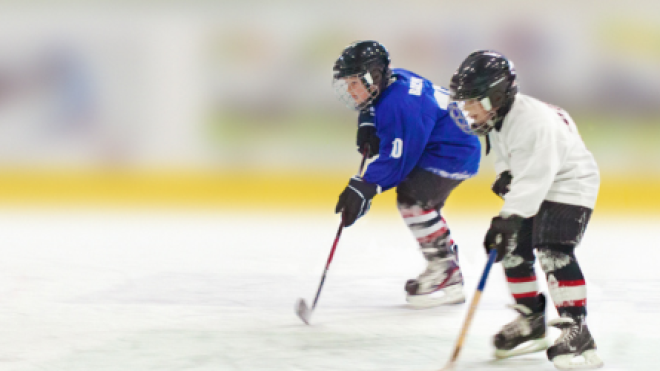 Two kids in hockey gear chasing a puck on an ice rink.