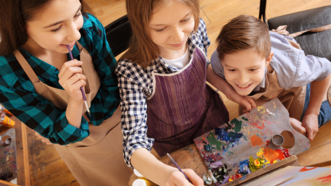 Three kids in aprons painting and smiling in an art class.