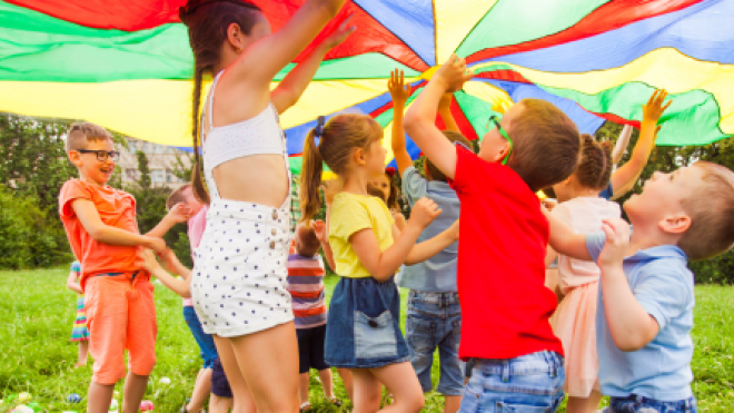 Children play under a colorful parachute in a grassy field.