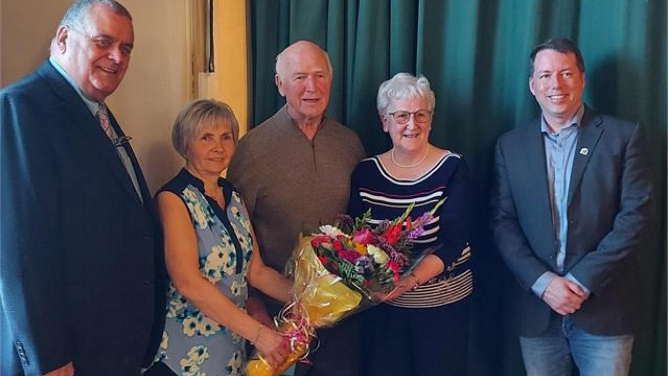 Five smiling adults standing, one holding a bouquet, in front of a green curtain.