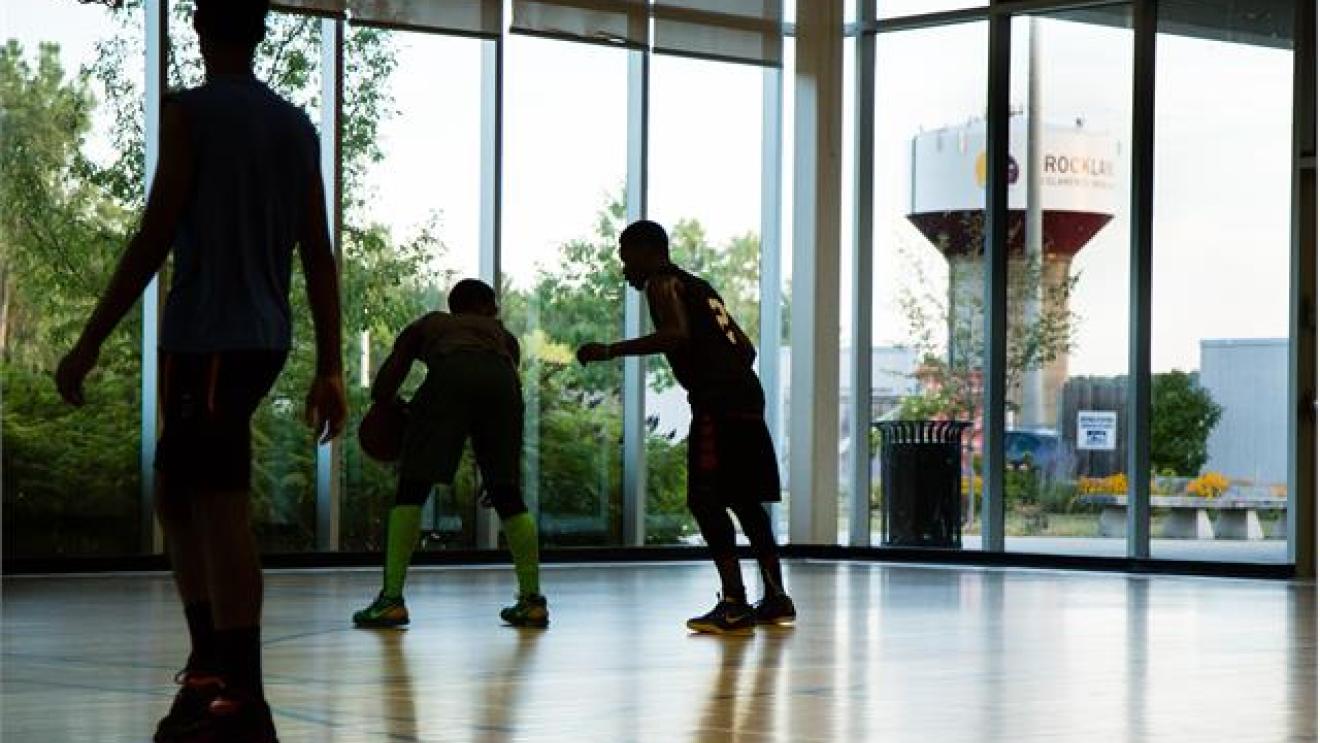 Three people playing basketball indoors, large windows in the background.