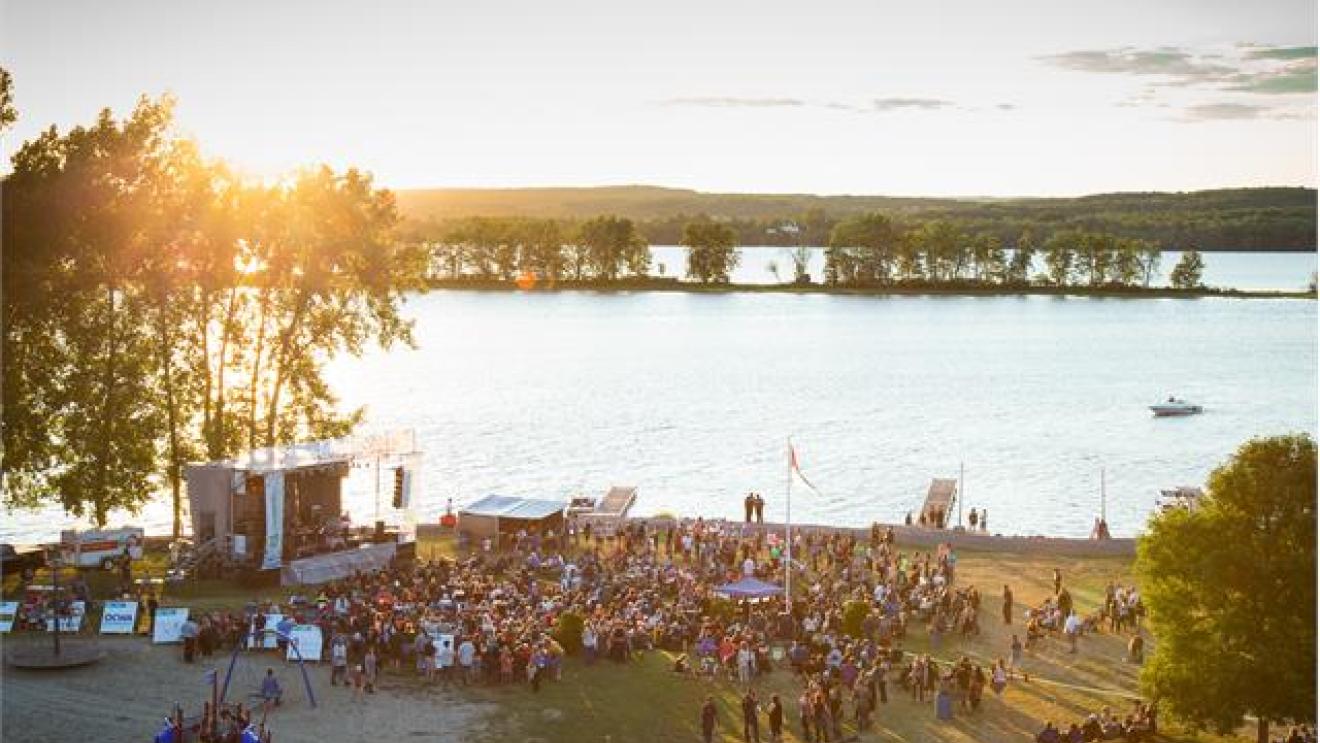 Outdoor concert by a lake at sunset, large crowd gathered near the stage.