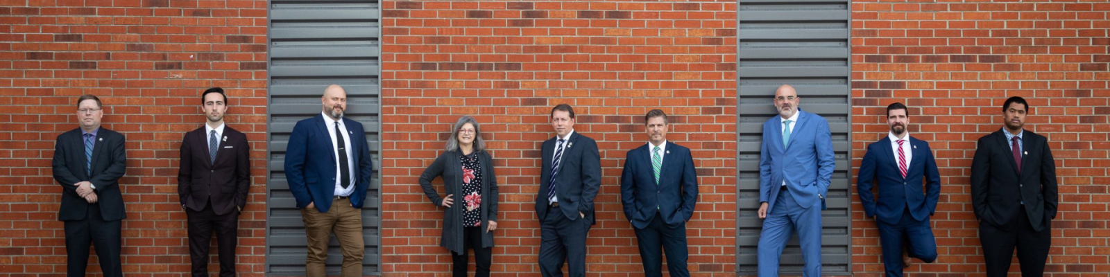 A group of nine people in formal attire against a brick wall.